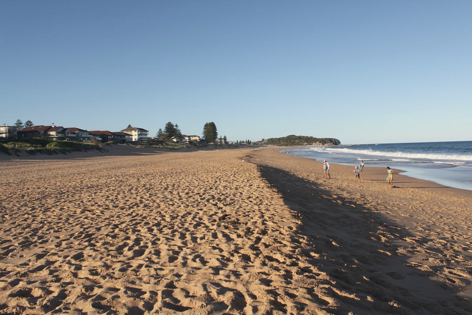 Warm late-afternoon light across the wide sands of Narrabeen Beach, with long shadows and a few people near the water.
