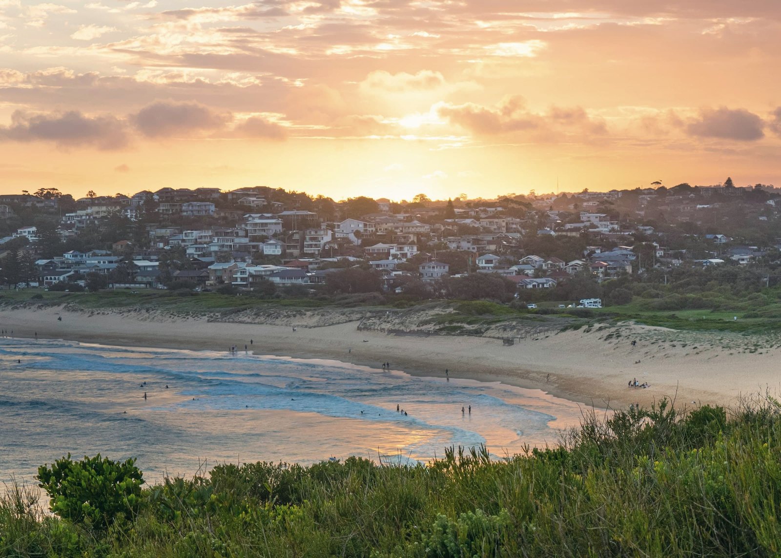 Houses on a hillside above a lagoon and beach at sunset, the sky streaked with orange cloud.