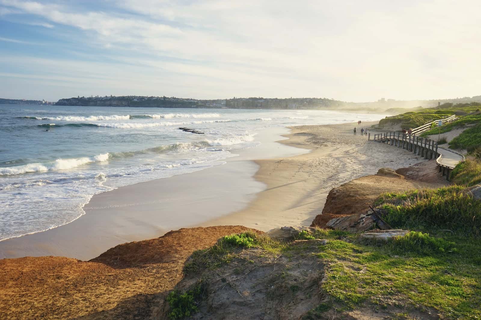 Morning light over a long beach and gentle surf, seen from a grassy headland with a boardwalk along its edge.